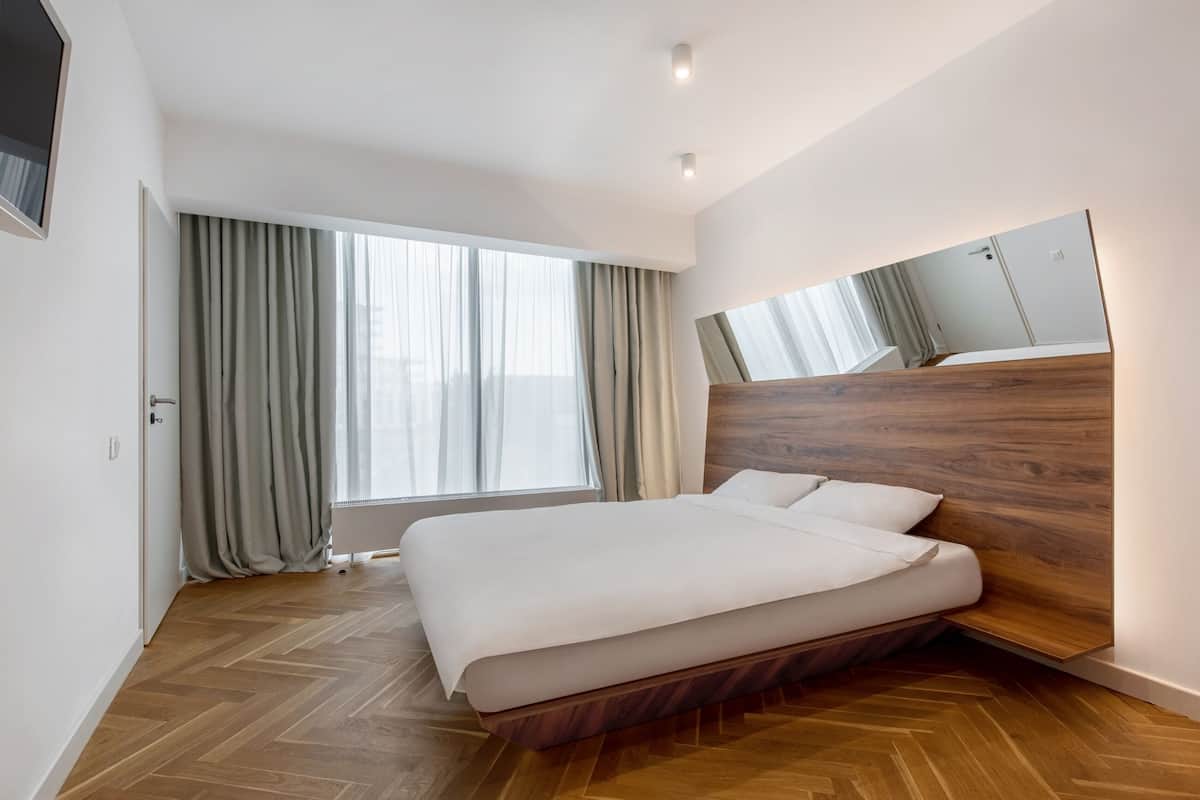 A modern bedroom with a wooden headboard, a large bed, a window with curtains, and a mirror on the wall.
