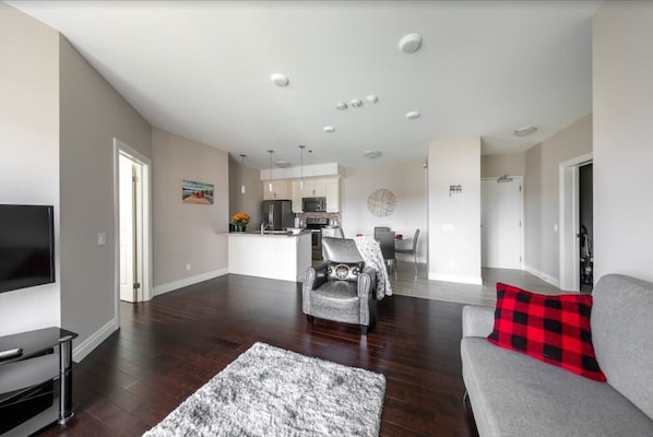 A modern living room with a grey sofa, a red and black plaid throw pillow, and a grey rug. There is a flat-screen TV on a stand, a black entertainment unit, and a dining area with a table and chairs. The kitchen is visible in the background with white cabinets and a microwave.