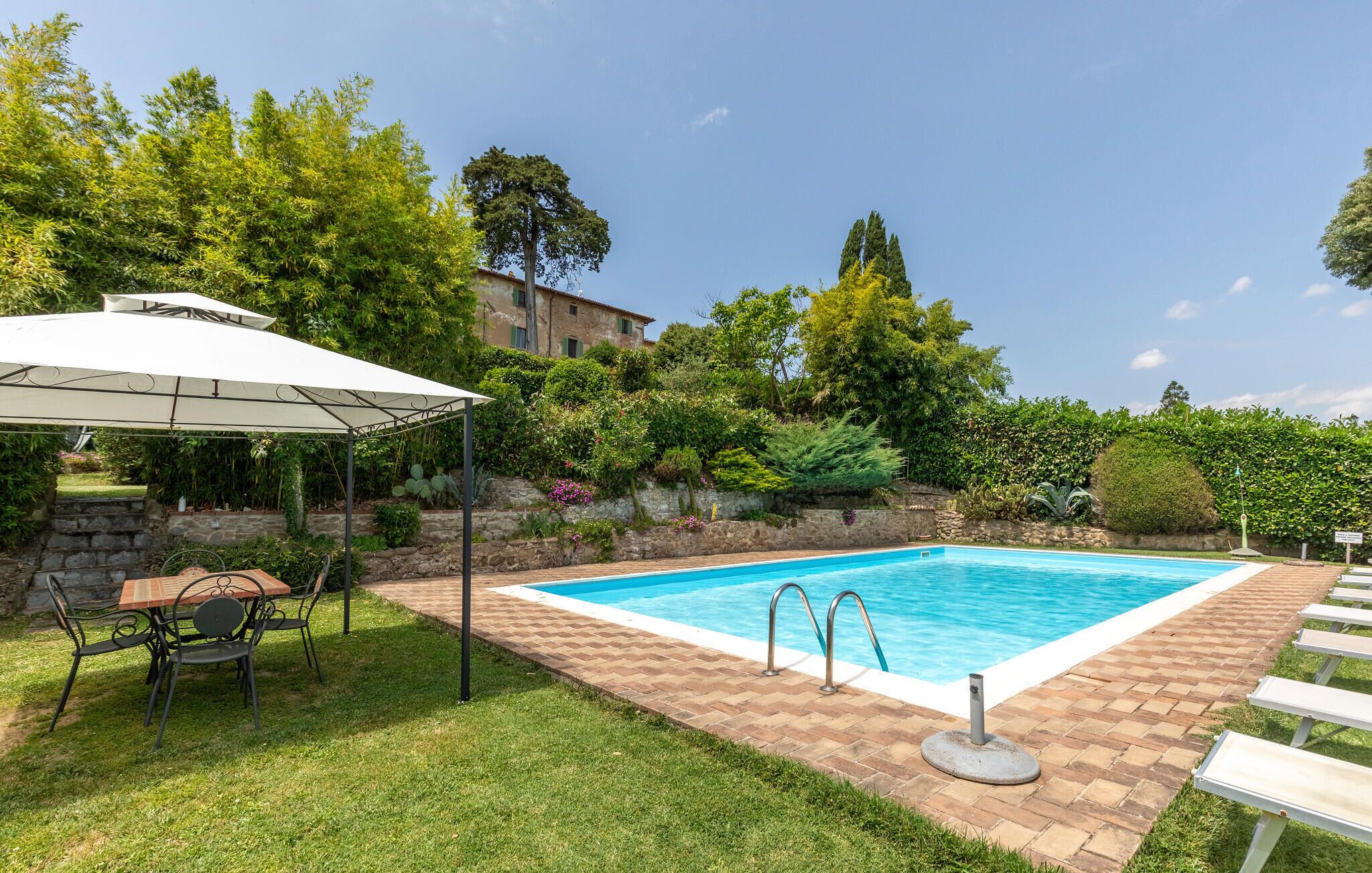 Ein Schwimmbecken mit Ziegelaußenbereich, eine überdachte Terrasse mit Tisch und Stühlen sowie eine Steinmauer mit Pflanzen.