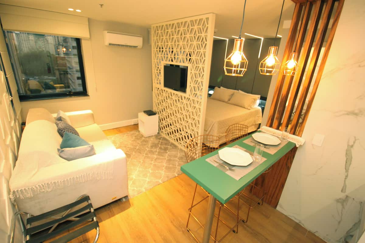 A compact hotel room with a sofa, a dining table set for two, a TV mounted on a decorative partition, and a view of the city through a window.