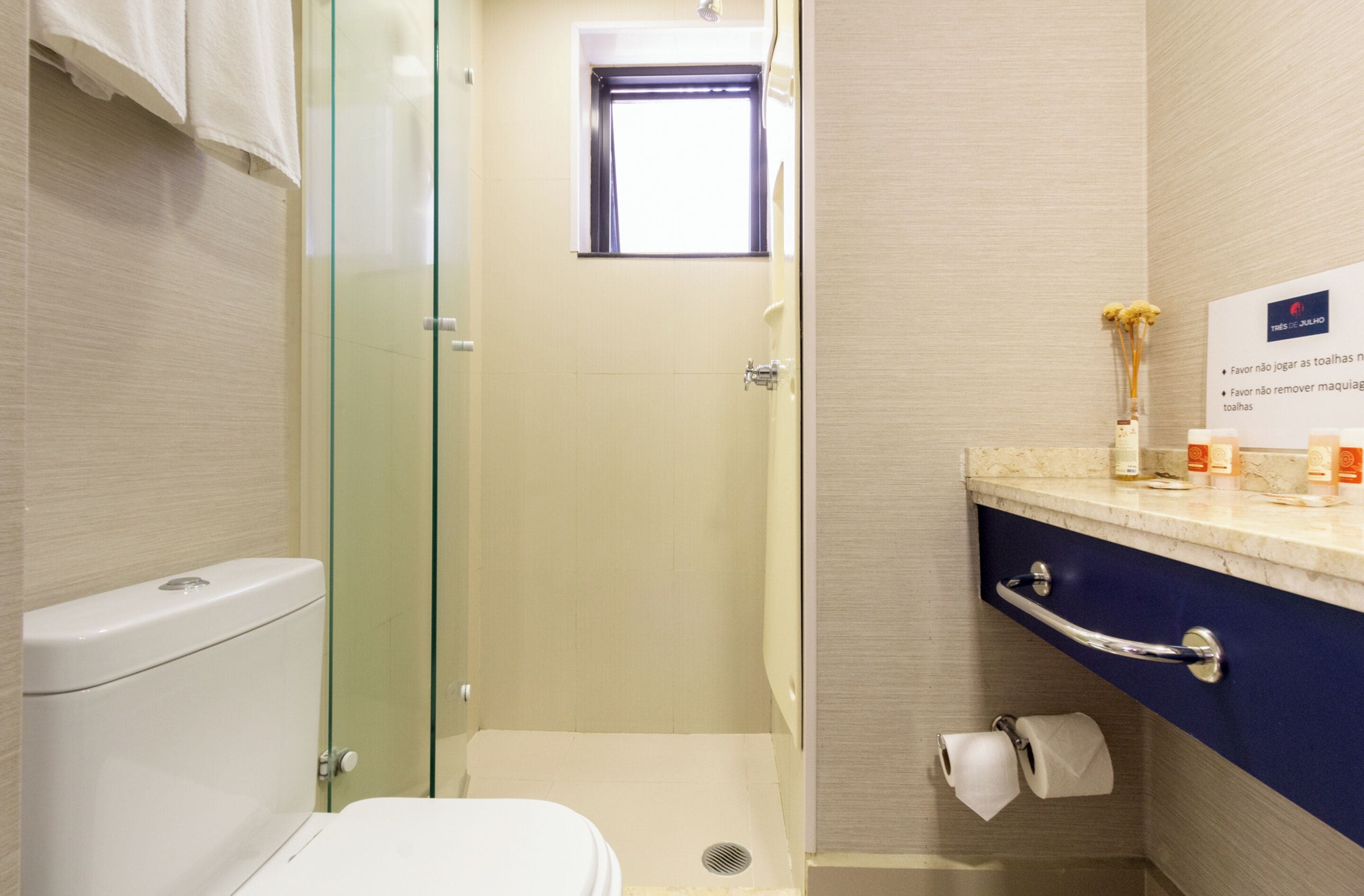 A bathroom with a glass shower enclosure, a toilet, a sink with a marble countertop, and a small window.