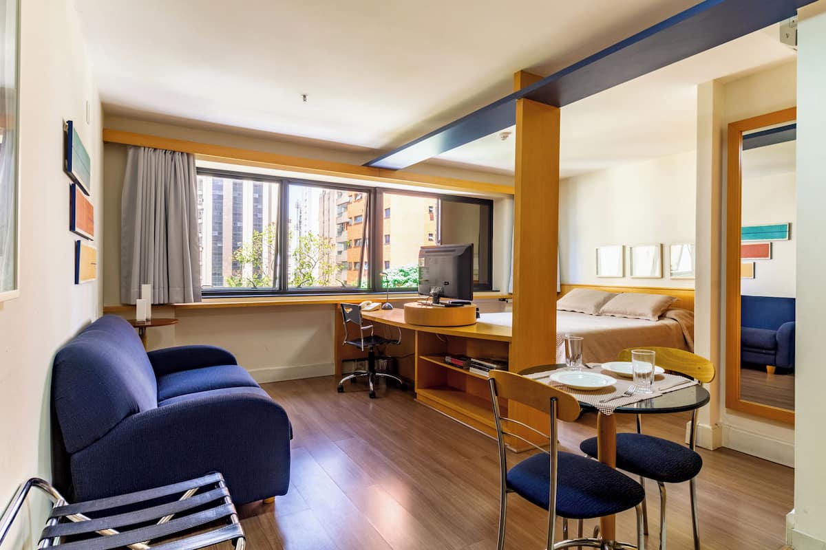 A compact hotel room with a sofa, a desk with a computer, a small dining table, and a bed.