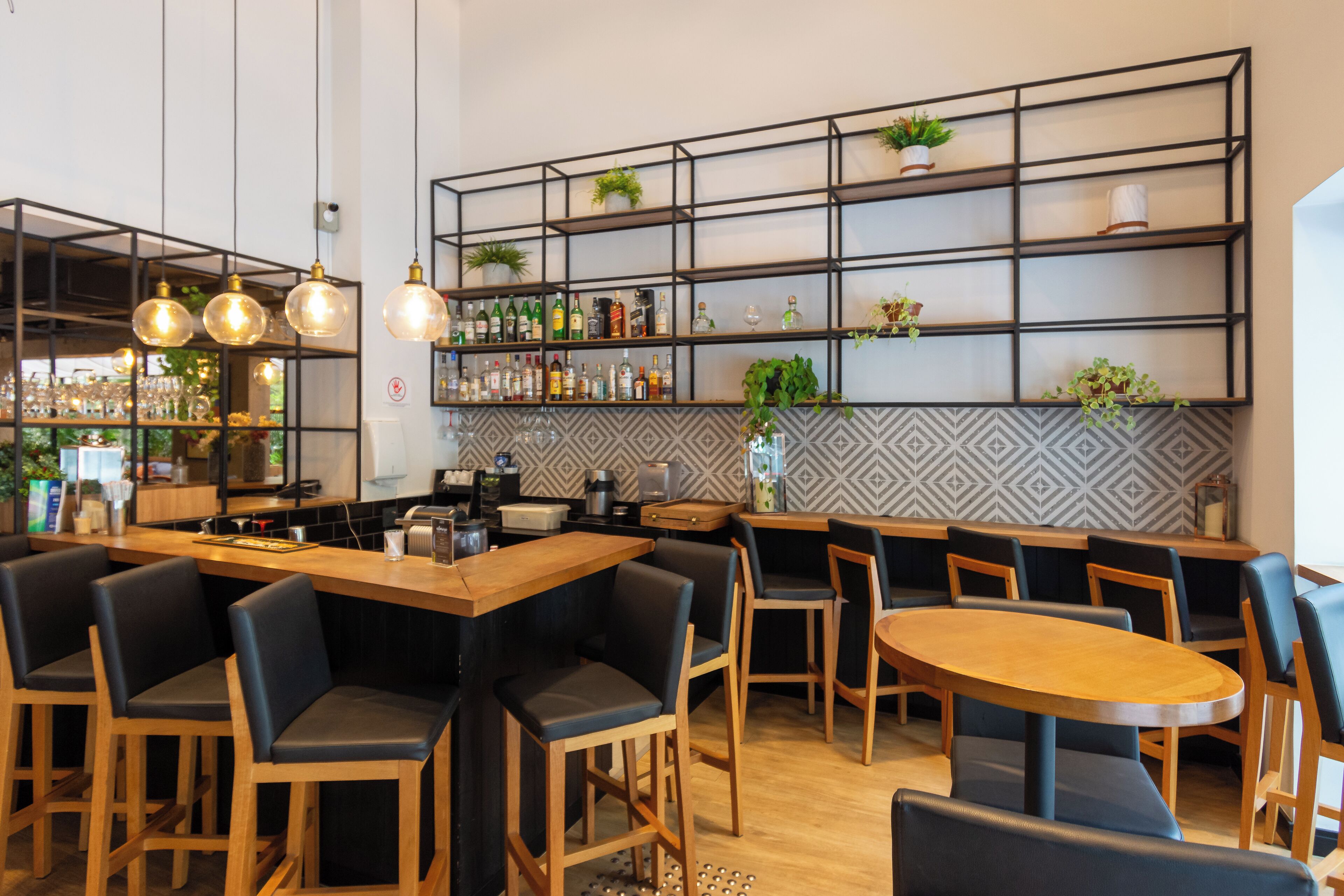 A modern bar area with wooden tables and chairs, shelves with bottles and plants, and hanging lights.