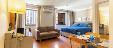 A hotel room with a bed, a sofa, a dining table with chairs, a desk with a lamp, and a telephone.