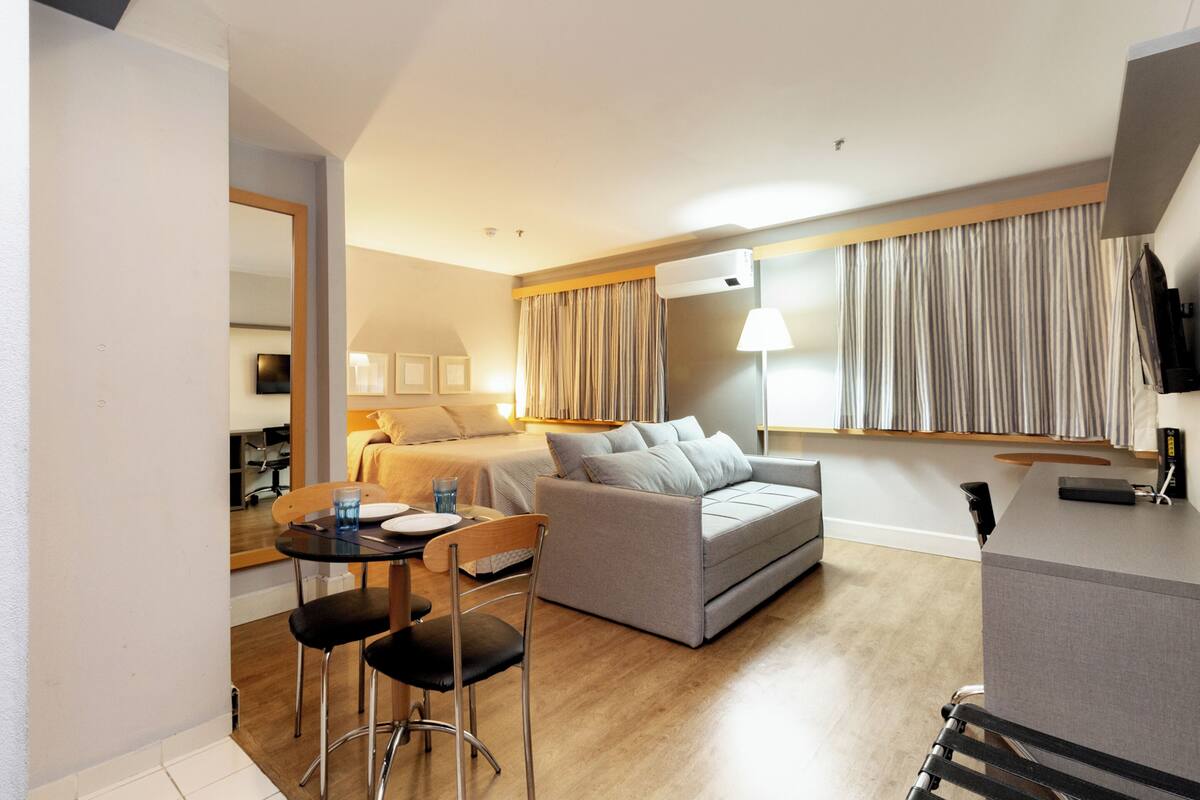 A modern hotel room with a sofa, a dining table with chairs, a TV, and a large window with curtains.