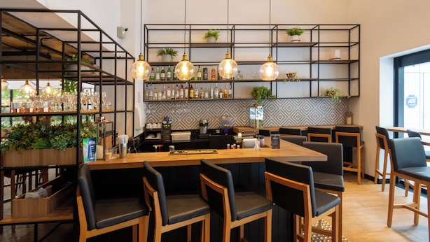 A modern bar area with wooden furniture, hanging pendant lights, and shelves stocked with bottles and glasses.
