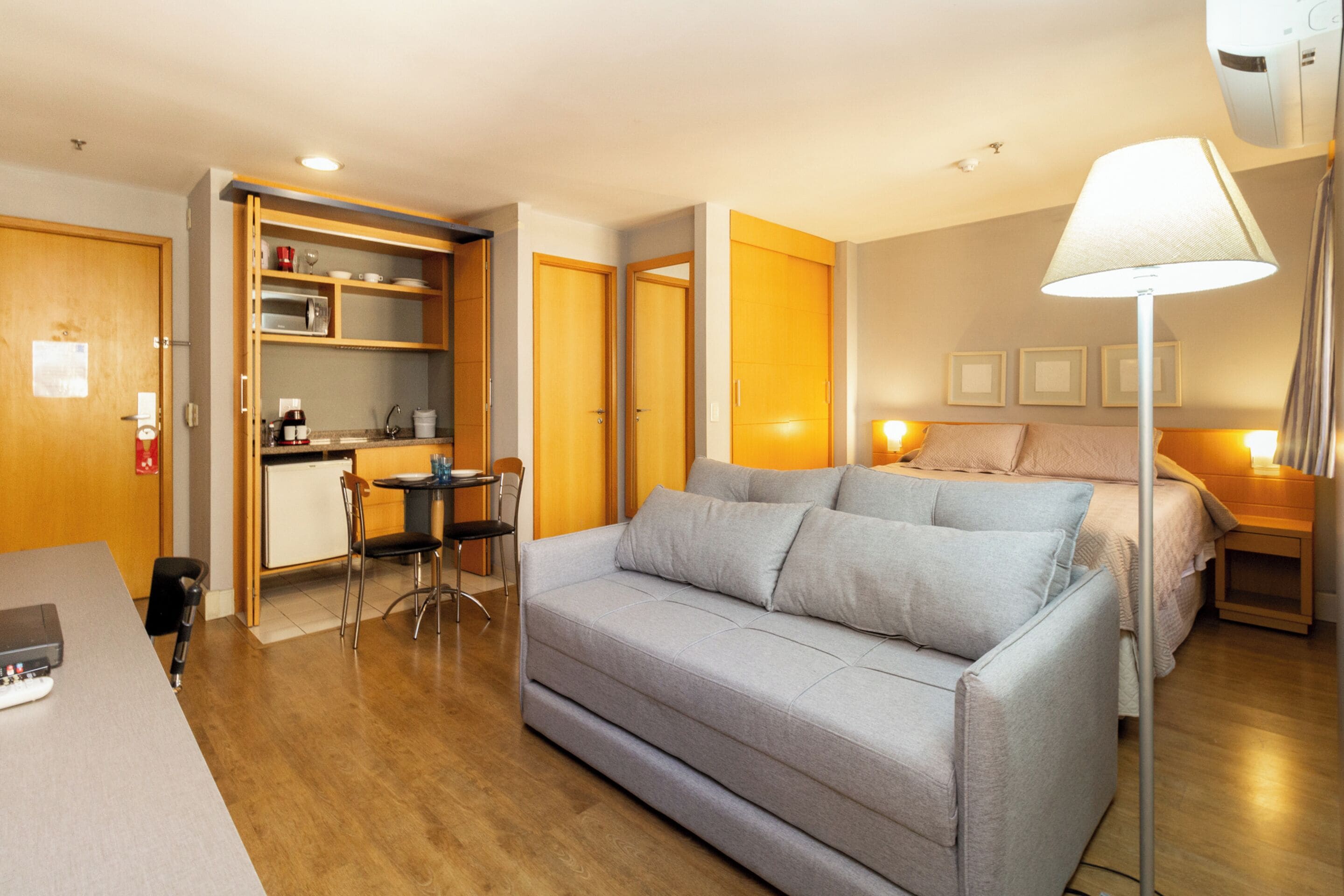 A compact hotel room with a grey sofa, a small kitchenette, a bed, and a bedside lamp.