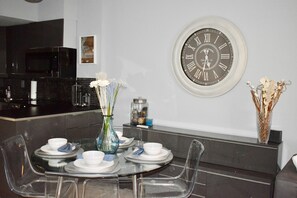 Comedor con una mesa redonda para cuatro, sillas transparentes, un reloj de pared y un jarrón con flores secas.