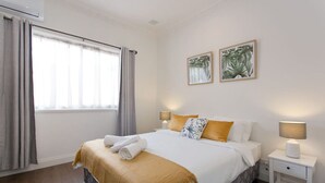 Ein ordentlich bezogenes Bett mit einer beigen Tagesdecke, zwei Kissen und einem gefalteten Handtuch an der Seite. Zwei Nachttischlampen und ein kleiner weißer Nachttisch mit Lampe sind ebenfalls vorhanden. Das Zimmer verfügt über ein großes Fenster mit Vorhängen und zwei gerahmte Bilder an der Wand.