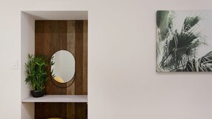 Ein Zimmer mit einer Holzwand, einem runden Spiegel, einem Topfpflanzen, einem gelben Hocker und einem Bilderrahmen mit Palmenmotiven.