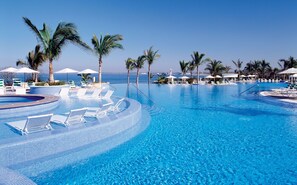 Ein großer, klarer, blauer Pool mit Liegestühlen und Sonnenschirmen, umgeben von Palmen und blauem Himmel.