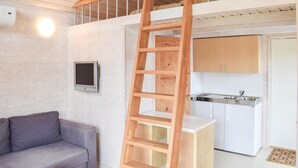A loft-style room with a wooden ladder, a gray sofa, a small table, a kitchen area with white cabinets, and a mounted TV. - Cozy home in Hagby with kitchen (Hagby)