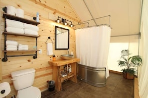 A wooden cabin bathroom with a toilet, sink, mirror, shelves with towels, and a shower curtain. - Zion Glamping Tent w/ A/C & Private Bathroom (Orderville)