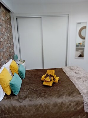 Een slaapkamer met een bruine bedspreide met patroon, een witte muur, een accentmuur van steen en een witte deur.