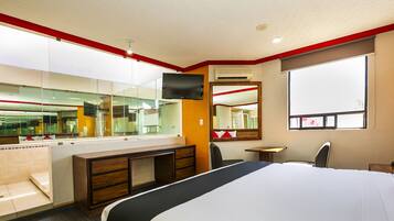 Habitación de hotel con una cama grande, un escritorio con silla, un espejo, un televisor y una ventana.