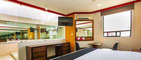 A hotel room with a large bed, a desk with a chair, a mirror, a television, and a window.