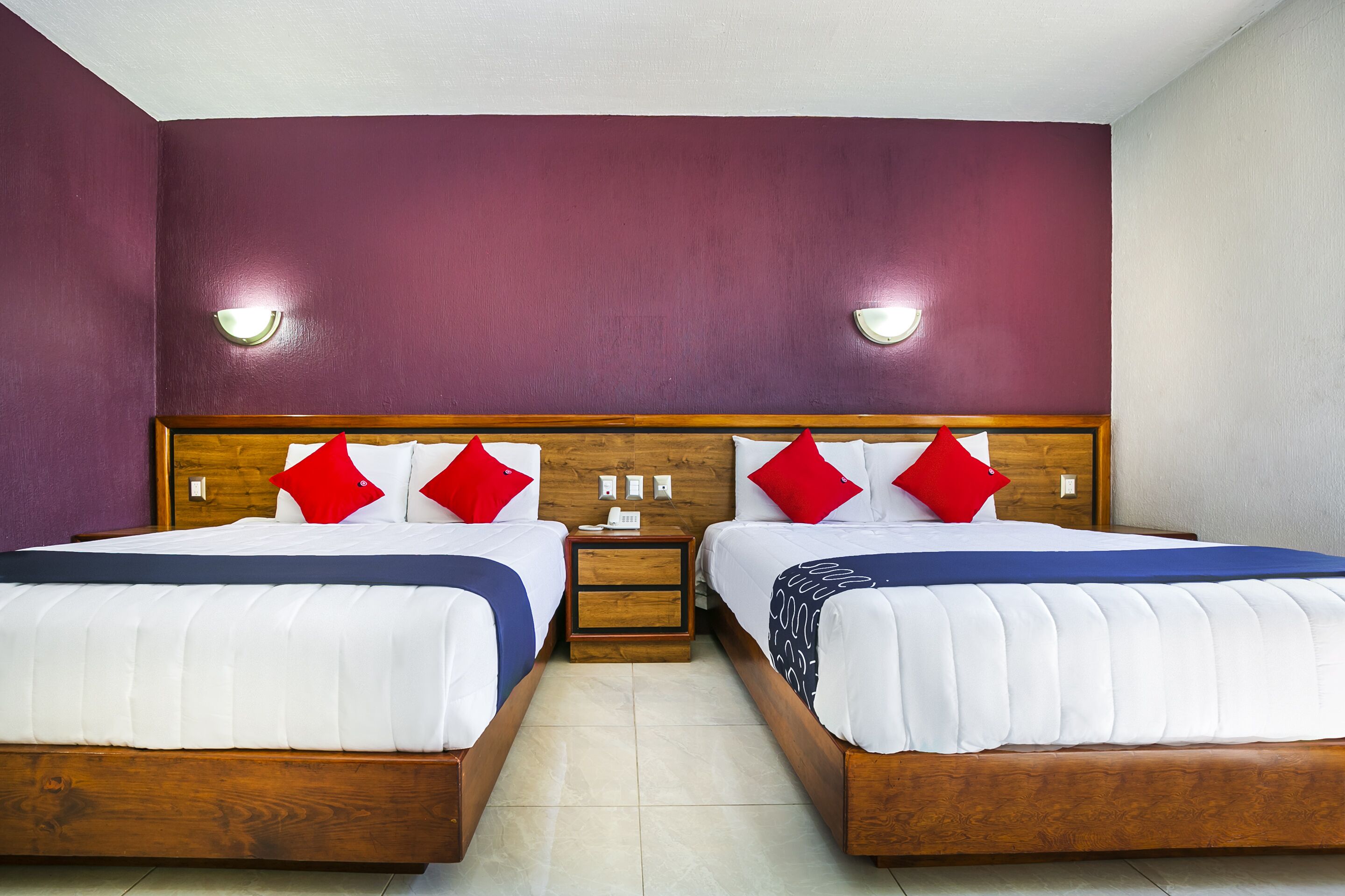 Una habitación doble con dos camas, cada una con ropa blanca y almohadas rojas, un cabecero de madera y una mesita de noche con teléfono y lámpara.