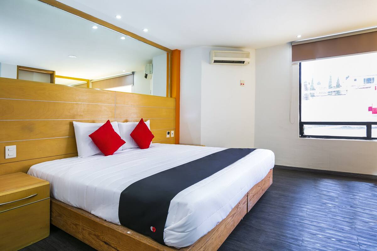 Una habitación de hotel moderna con cabecera de madera, una cama grande con ropa de cama blanca y almohadas rojas, una mesita de noche de madera, una ventana con vistas y un equipo de aire acondicionado en la pared.