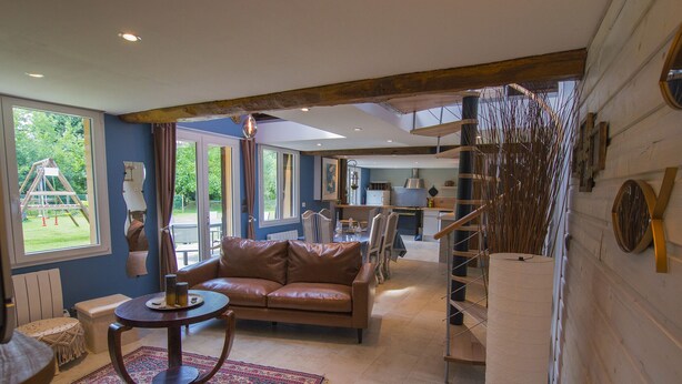 A spacious living area with a leather sofa, a round wooden table, and a patterned rug. A staircase leads to an upper level, and a large window offers a view of a garden. - THE PIRATES FARM (MANNEVILLE la raoult)