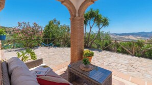 A patio with a wicker sofa, a small table with a plant, and a view of hills and trees. - Villa Gema Antequera - La Joya by Ruralidays (Málaga)