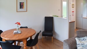 A dining area with a wooden table and chairs, a black chair, a framed picture on the wall, a window with a view, and a kitchen area with a refrigerator. - 2 bedroom accommodation in Mount Hawke, near Redruth (Mount Hawke, near Redruth)