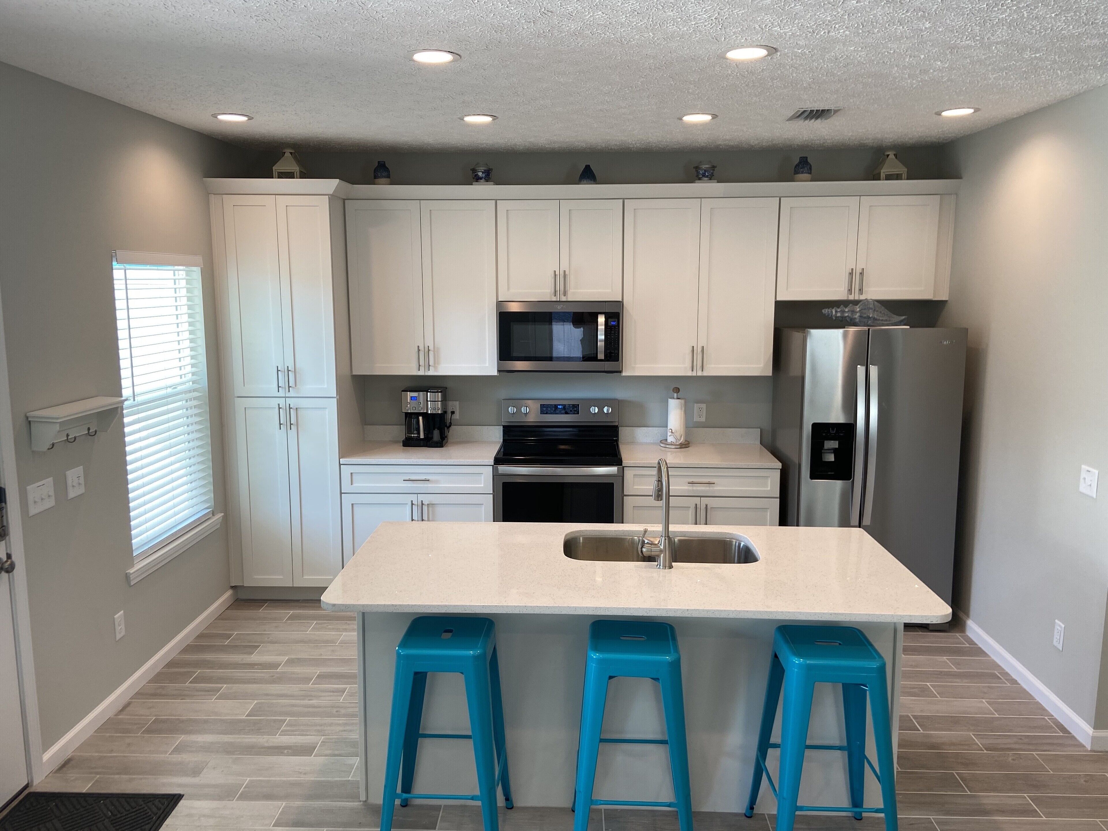 A modern kitchen with white cabinets, a stainless steel refrigerator, a microwave, and a coffee maker. There are three blue barstools and a sink with a faucet.
