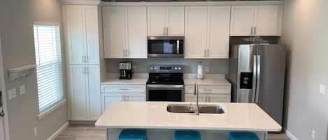 A modern kitchen with white cabinets, a stainless steel refrigerator, a microwave, and a coffee maker. There are three blue barstools and a sink with a faucet.