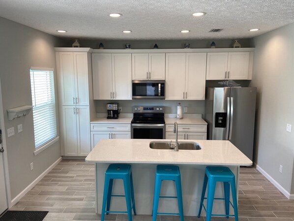 A modern kitchen with white cabinets, a stainless steel refrigerator, a microwave, and a coffee maker. There are three blue barstools and a sink with a faucet.