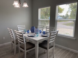 A dining area with a white table set for four, surrounded by white chairs with dark cushions. There are blue glass vases on the table, and the room has a gray wall and wooden flooring.