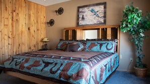 Une chambre avec un mur en bois, un lit avec une literie à motifs, une lampe fixée au mur, un tableau et une plante en pot.