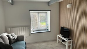 A modern living room with a sofa, a TV on a white stand, a window with blinds, and a radiator. - Linhay, character 1 bed 19th century cottage, peaceful location (Bude)