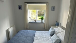 A bedroom with a bed, a window with curtains, a bedside table with a lamp, and a wall-mounted air conditioner. - Linhay, character 1 bed 19th century cottage, peaceful location (Bude)
