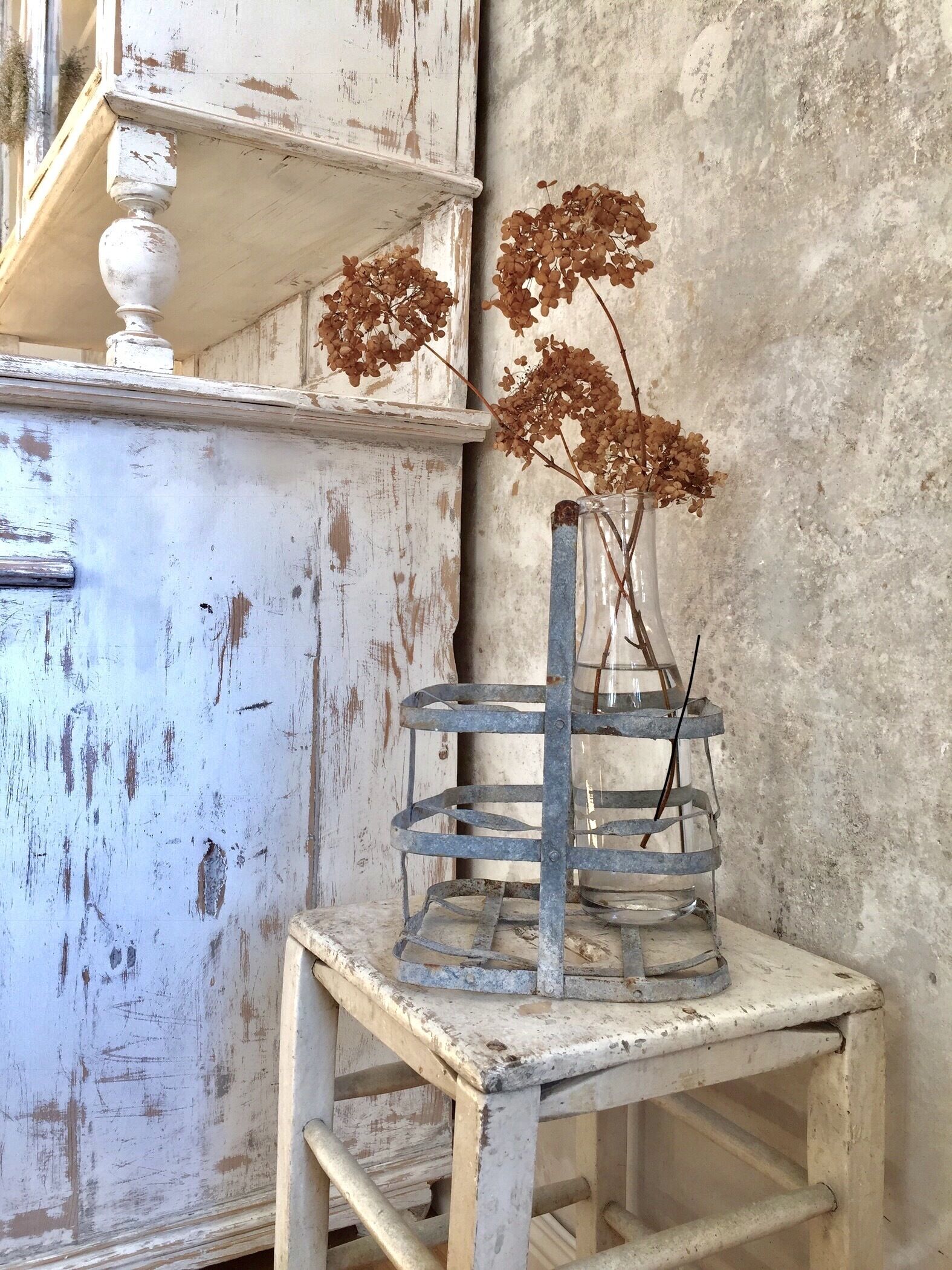 A rustic, weathered white dresser with a decorative top and a metal shelf unit beside it.