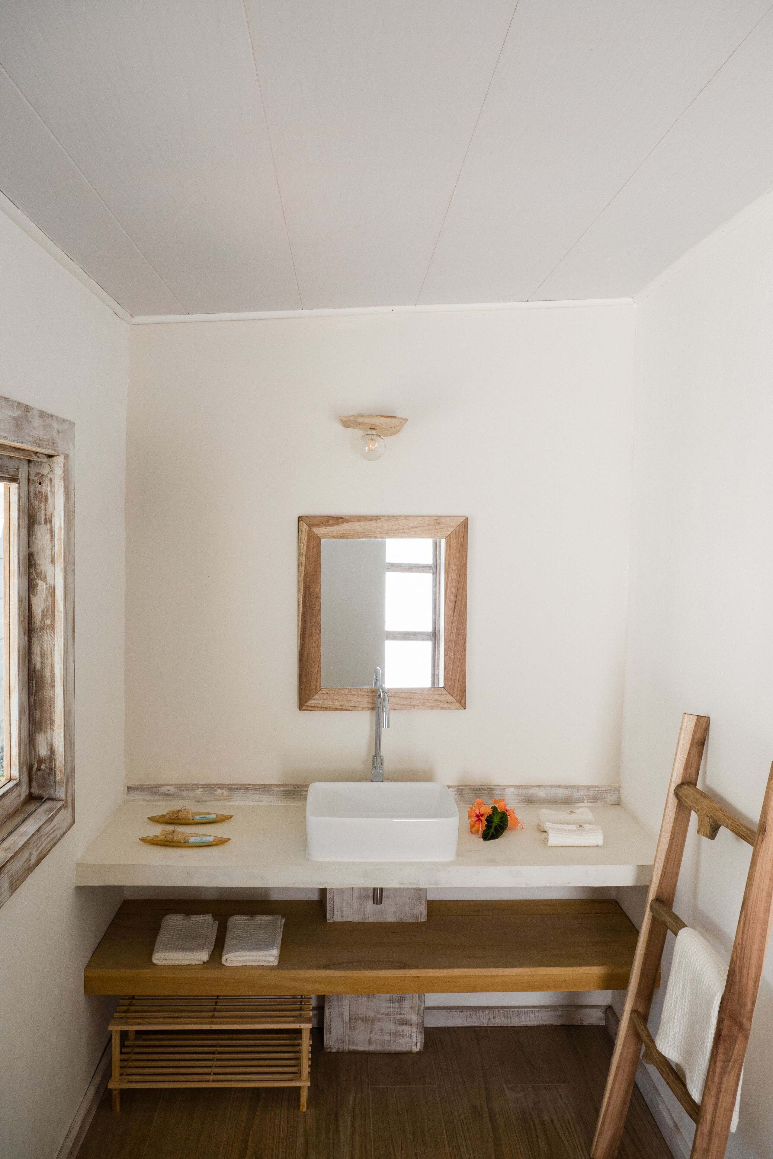 A bathroom with a wooden bench, a sink, a mirror, and a window with a wooden frame.