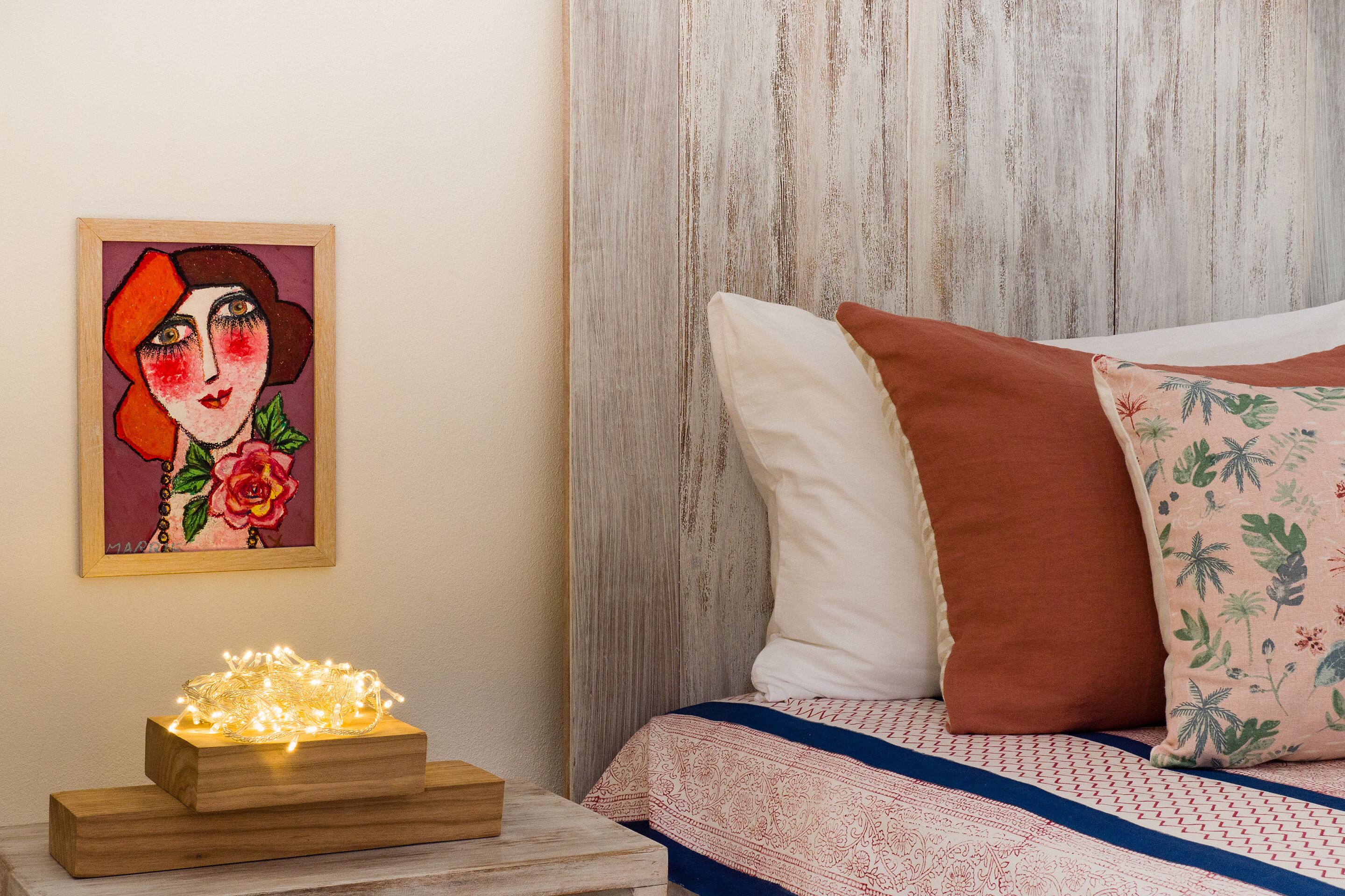 Un dormitorio con cabecera de madera, cama con ropa de cama estampada, mesita de noche con luces y un cuadro en la pared.
