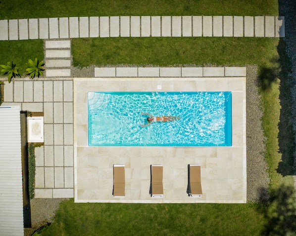 Eine Luftaufnahme eines rechteckigen Schwimmbeckens, in dem eine Person schwimmt, umgeben von einer gepflasterten Liegefläche, zwei Liegestühlen und einer kleinen Gartenanlage mit Pflanzen.