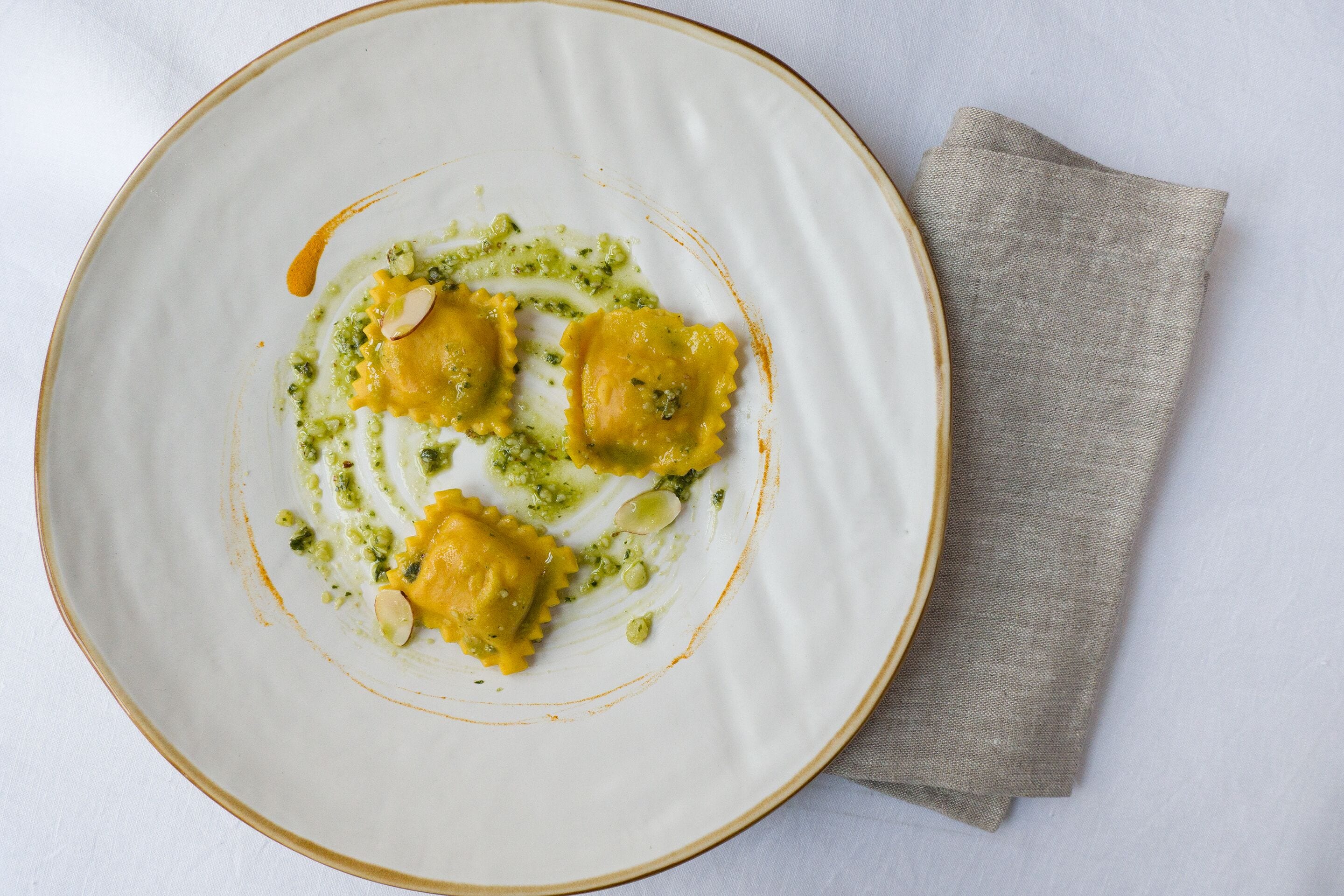 A plate of ravioli with pesto sauce and almonds, placed on a white plate with a beige napkin beside it.