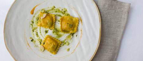A plate of ravioli with pesto sauce and almonds, placed on a white plate with a beige napkin beside it.