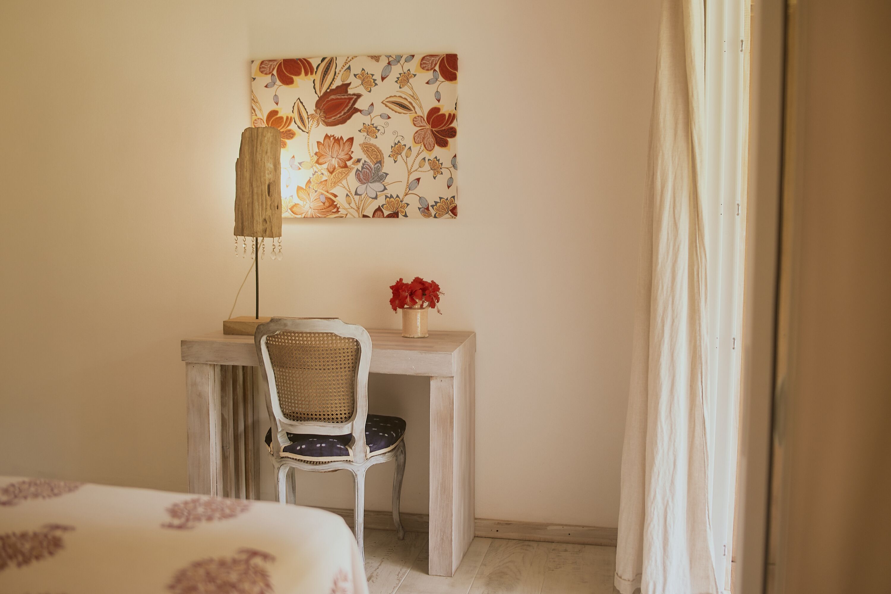 A room with a bed, a desk with a chair, a wall-mounted lamp, and a floral painting.