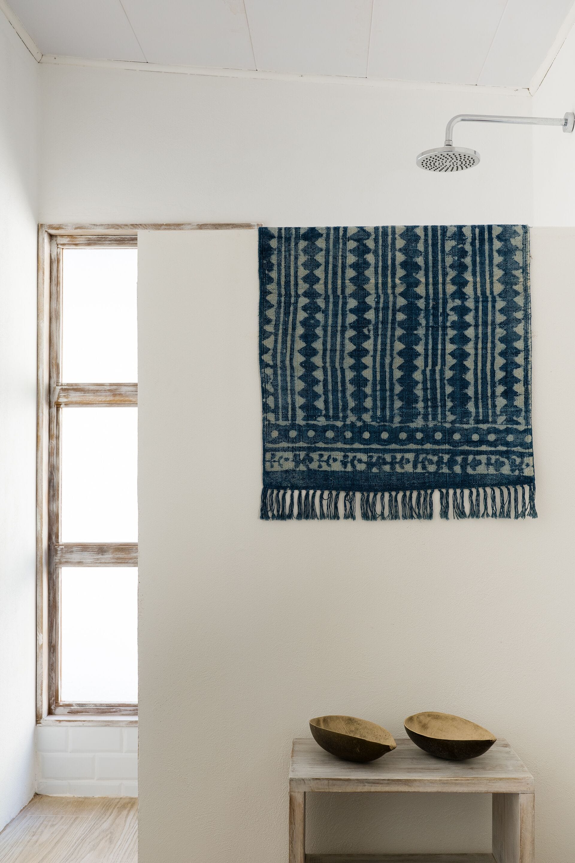 Une pièce dans laquelle on peut voir un mur orné d’un tissu à motifs bleus et blancs, une petite table en bois sur laquelle reposent deux bols, et une pomme de douche.