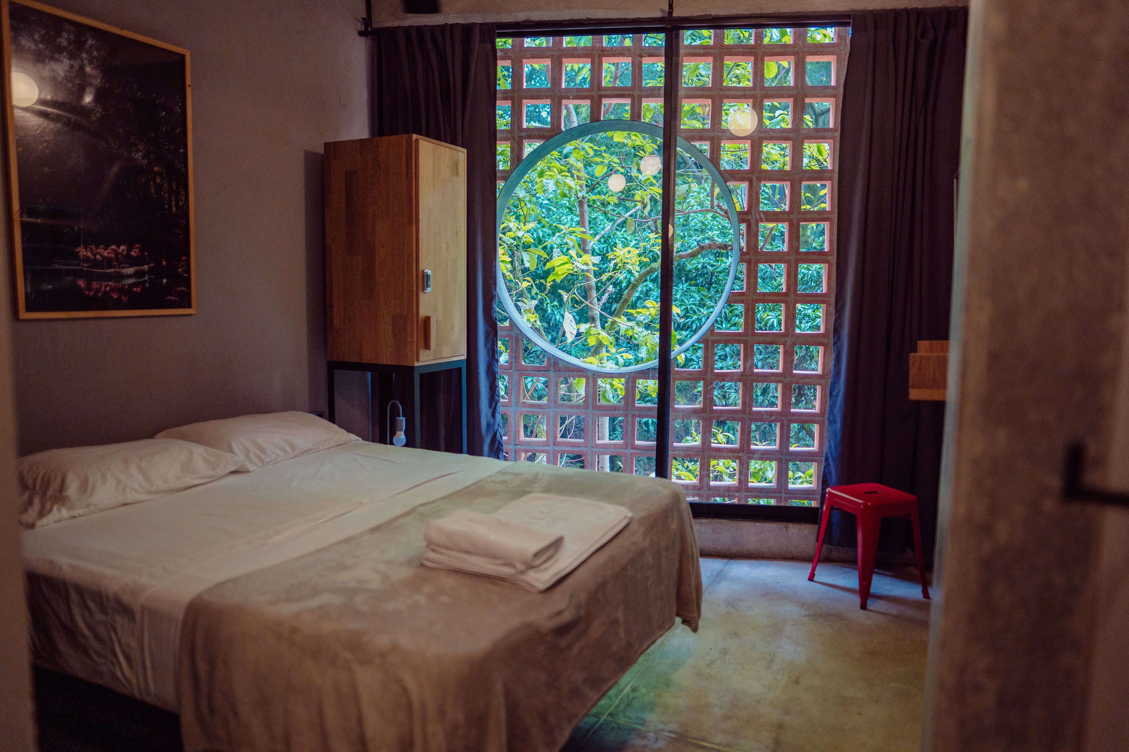 A bedroom with a bed, a wooden cabinet, a window with a circular view, and a red stool.