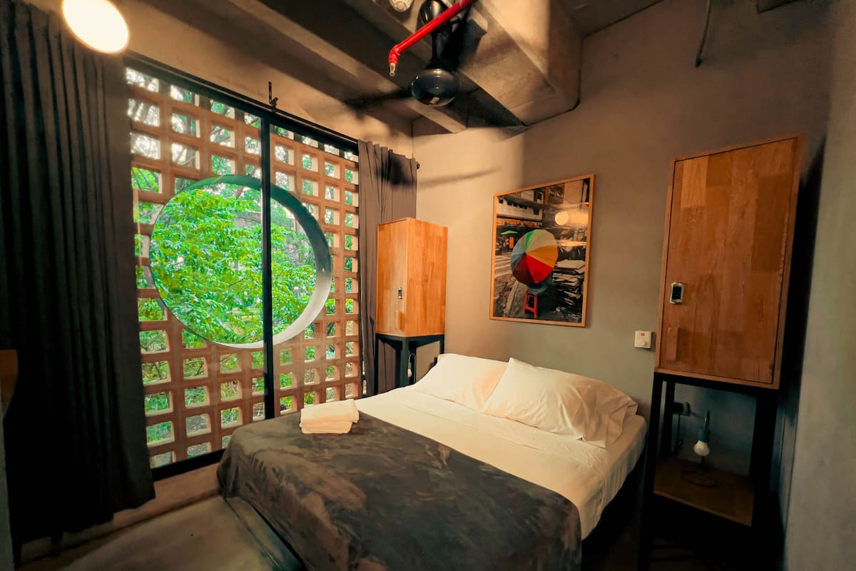 A bedroom with a bed, a window with a view of greenery, a wooden cabinet, and a painting on the wall.