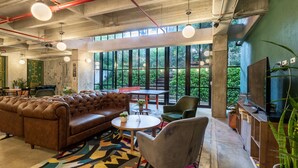 A spacious lounge area with a leather sofa, a round table, and a television. Large windows provide a view of greenery outside. - Viajero Hostel Medellin (Medellín)