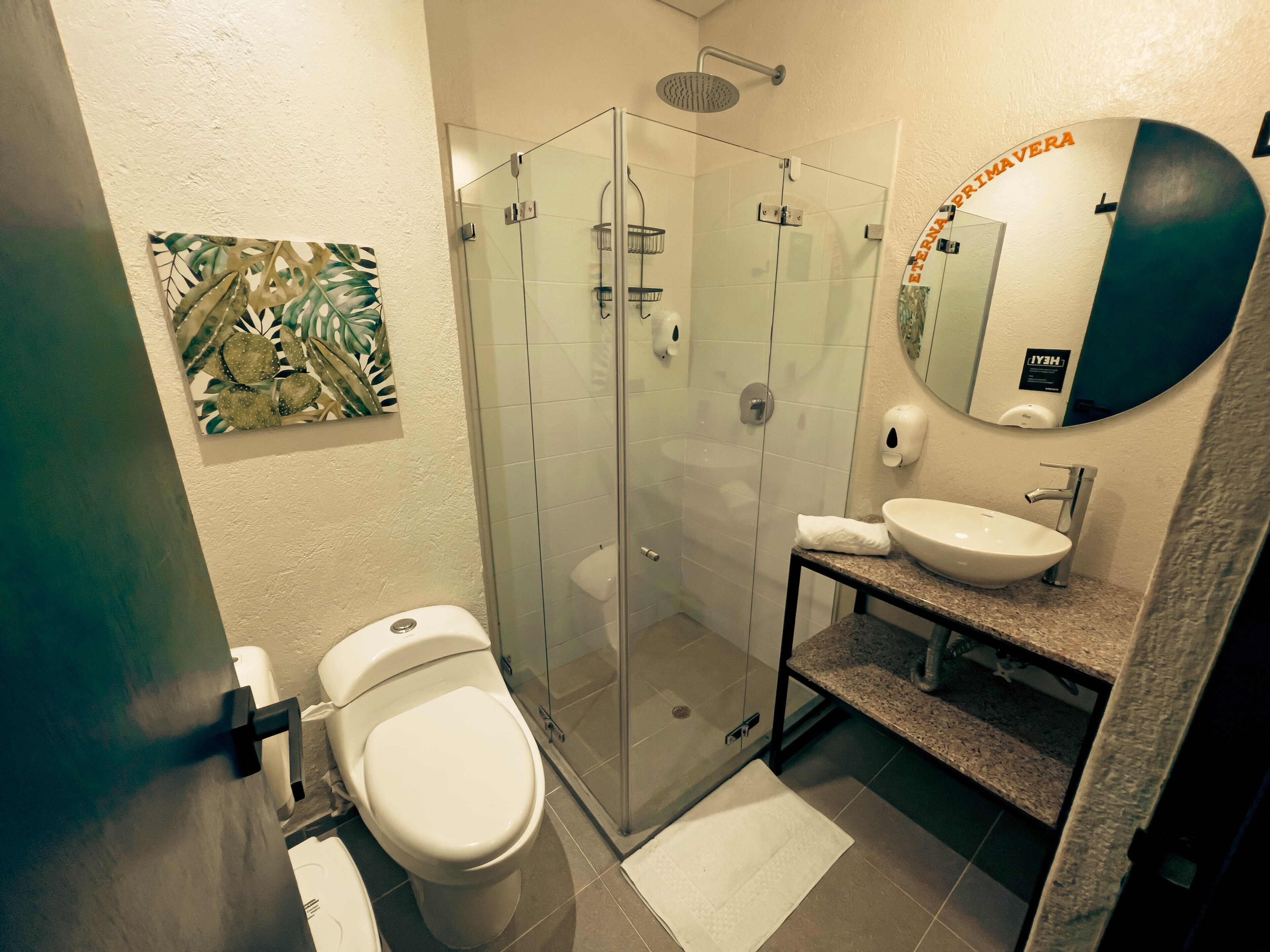 A modern bathroom with a glass-enclosed shower, a white toilet, a sink with a mirror, and a decorative leaf wall art piece.