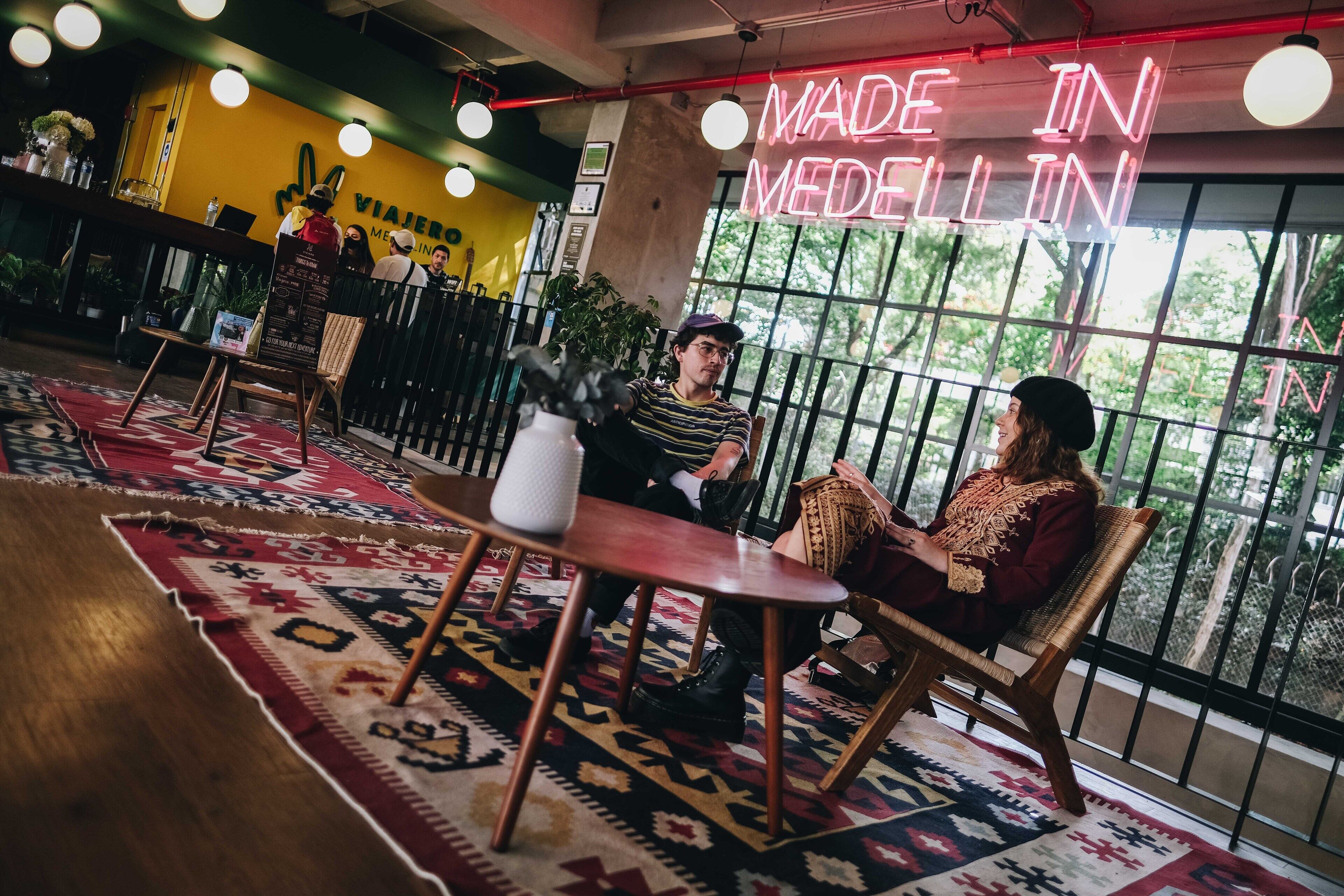 A cozy interior with a 'Made in Medellín' neon sign, wooden furniture, and large windows offering a view of greenery.