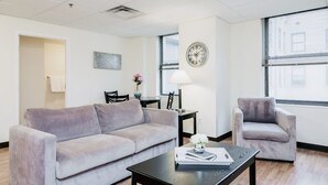 A modern living room with a sofa, armchair, and coffee table. There is a dining table with chairs, a lamp, and a clock on the wall.