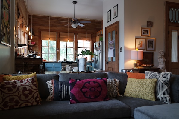 A cozy living room with a gray sofa, various patterned cushions, a ceiling fan, and a kitchen in the background.