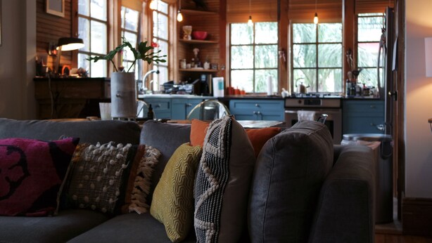 A cozy living room with a gray sofa adorned with various cushions, a wooden dining table, and a kitchen area with blue cabinets and a window overlooking greenery. - open for summer 2020 (New Orleans)