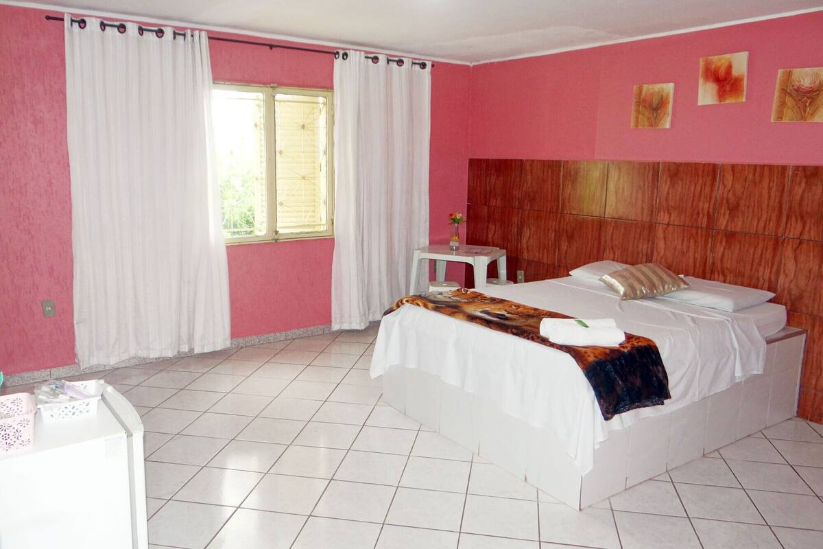 A hotel room with a bed, a small table, a window with curtains, and a pink wall.
