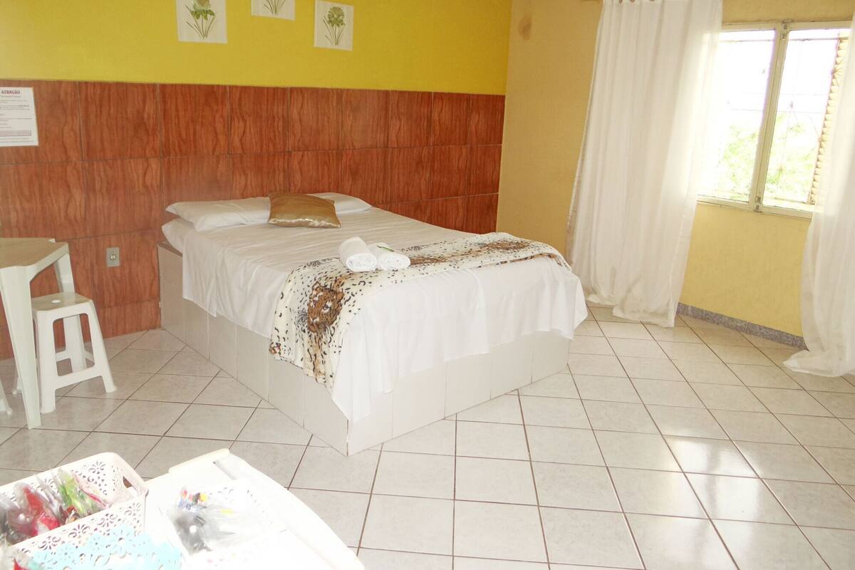 A hotel room with a bed, a small table with a chair, a window with curtains, and a wall with three framed pictures.